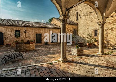 Mittelalterlicher Innenhof in der Stadt Recanati. Portico mit gewölbten Gewölben und Säulen mit dekorierten Hauptstädten und einem Brunnen. Ziegelboden in verschiedenen Farben Stockfoto