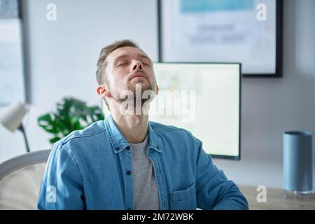 Müde Software-Entwickler-Konzept: Attraktiver IT-Support männlich im Jeansshirt, der mit geschlossenen Augen aufblickt. Bärtiger weißer, männlicher, freiberuflicher Software-Ingenieur am Arbeitsplatz im Büro. Hochwertige Bilder Stockfoto