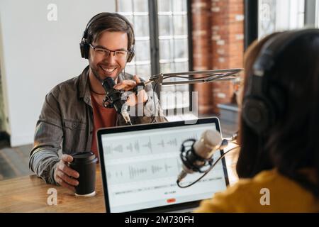 Fröhlicher junger Mann mit Kopfhörern, der in die Kamera schaut, lächelt, in einem Podcasting-Studio im Mikrofon spricht, während er ein Interview hat. Radiomoderator interviewt berühmte Bloggerin für eine Online-Sendung auf ihrem Kanal Stockfoto