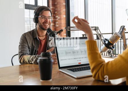 Radio-Podcast-Konzept. Fröhlicher männlicher Radiomitgast, der eine Gastgeberin ansieht, Kopfhörer trägt und eine Frage für einen Audio-Podcast im Studio beantwortet. Frau interviewt berühmte Influencer für Kanalinhalte Stockfoto