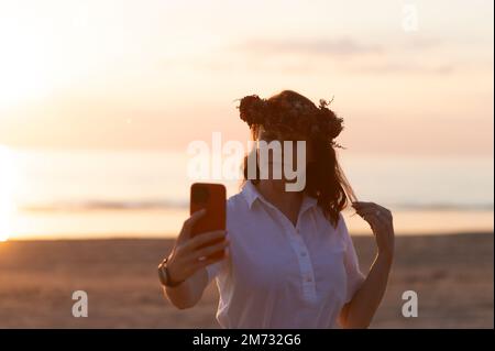 Junge Frau, die Selfie über das Smartphone nimmt, während sie während des Sonnenuntergangs am Strand steht Stockfoto