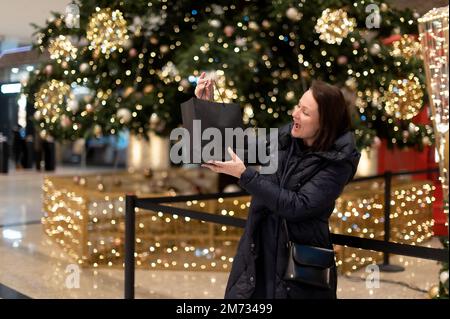 Glückliche Frau, die eine Einkaufstasche auf dem Hintergrund eines Weihnachtsbaums mit hellen Lichtern hält. Stockfoto