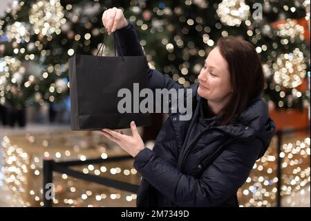 Glückliche Frau, die eine Einkaufstasche auf dem Hintergrund eines Weihnachtsbaums mit hellen Lichtern hält. Stockfoto