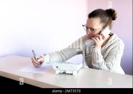 Eine Frau mittleren Alters spricht am Telefon und schreibt sich am Arbeitsplatz Notizen Stockfoto