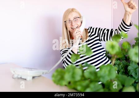 Glückliche Frau mittleren Alters, die am Arbeitsplatz über ein Festnetztelefon spricht und Daumen nach oben zeigt Stockfoto