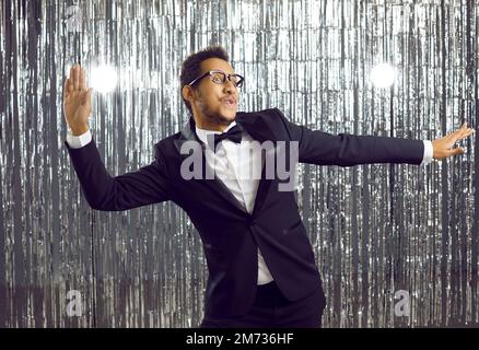 Positiver junger afroamerikanischer Mann in elegantem Anzug, der Spaß beim Tanzen auf der Party hat, auf glänzendem Hintergrund. Stockfoto