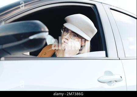 Eine traurige Frau mittleren Alters mit Brille und Hut sitzt in einem Auto und beobachtet ihr Gesicht im Rückspiegel Stockfoto