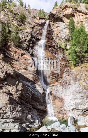 Lillaz-Wasserfall (Cascate di Lillaz) und Granitfelsen mit immergrünen Bäumen unter blauem Himmel mit weißen Wolken. Aosta-Tal, Italien, vertikaler Schuss Stockfoto