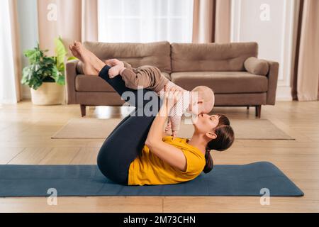 Sport mit dem Baby. Die mutter küsst ihren kleinen Sohn, während sie ihn an den Beinen hält und postpartale Erholungsübungen auf der Fitnessmatte zu Hause macht. Sportliche Frau, die zusammen mit ihrem kleinen Baby trainiert Stockfoto