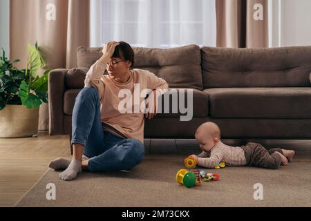 Junge müde Frau, die an postnataler Depression leidet, ihren Kopf mit der Hand hält, auf dem Boden im Wohnzimmer neben ihrem Kleinkinder sitzt und mit Spielzeug spielt. Neue mutter müde von Mutterschaftsschwierigkeiten Stockfoto