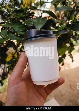 Halten Sie eine weiße Tasse Kaffee zum Mitnehmen mit grünen tropischen Blättern im Hintergrund in der Hand Stockfoto