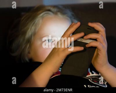 Ein Teenager liegt im Bett und hält sein Smartphone, bevor er einschläft. Das Smartphone-Display beleuchtet das Gesicht des Jungen. Kaukasische ethnische Zugehörigkeit. Stockfoto