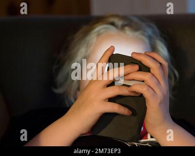 Ein Teenager liegt im Bett und hält sein Smartphone, bevor er einschläft. Das Smartphone-Display beleuchtet das Gesicht des Jungen. Kaukasische ethnische Zugehörigkeit. Stockfoto