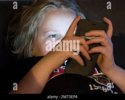 Ein Teenager liegt im Bett und hält sein Smartphone, bevor er einschläft. Das Smartphone-Display beleuchtet das Gesicht des Jungen. Kaukasische ethnische Zugehörigkeit. Stockfoto