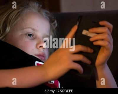 Ein Teenager liegt im Bett und hält sein Smartphone, bevor er einschläft. Das Smartphone-Display beleuchtet das Gesicht des Jungen. Kaukasische ethnische Zugehörigkeit. Stockfoto