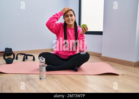 Junge hispanische Frau, die auf Yoga-Matte sitzt und Apfel isst, gestresst und frustriert von Hand auf Kopf, überrascht und wütend im Gesicht Stockfoto