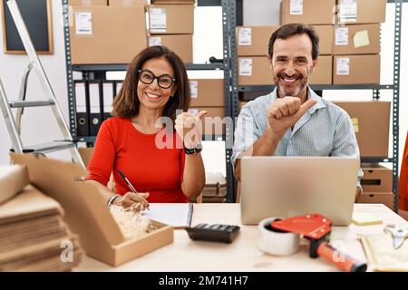 Ein Paar mittleren Alters, das im E-Commerce für kleine Unternehmen arbeitet und mit der Hand und den Daumen nach hinten zeigt und selbstbewusst lächelt Stockfoto