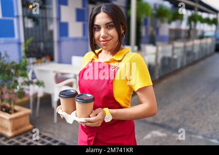 Junge schöne arabische Kellnerin lächelt selbstbewusst und hält Kaffee zum Mitnehmen auf der Terrasse des Cafés Stockfoto