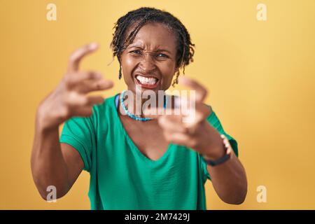 Afrikanische Frau mit Dreadlocks auf gelbem Hintergrund, die frustriert vor Wut schreit, Hände versucht zu erwürgen, schreit wütend Stockfoto