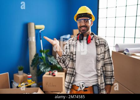 Junger lateinamerikanischer Mann mit Bart, der bei der Renovierung zu Hause arbeitet, lächelt fröhlich und zeigt mit der Handfläche in die Kamera. Stockfoto
