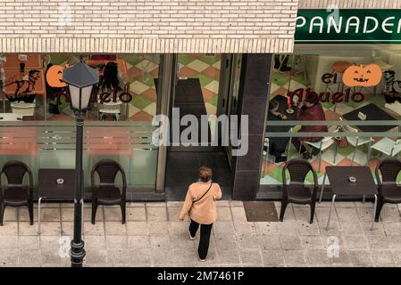 Luftaufnahme einer Dame in einem beigen Mantel auf der Straße, um eine Cafeteria-Bäckerei zu betreten, in der Leute sitzen und in das Fensterglas schauen. Stockfoto
