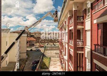 Arbeiter auf einem Kran mit Hebekorb repariert die Wand des Gebäudes. Der Baumeister in der hydraulischen Hebebühne reinigt die Dachrinne der Fassade. Stockfoto