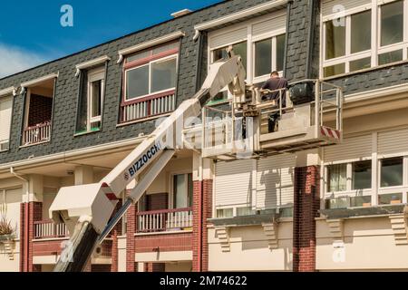 Arbeiter auf einem Kran mit Hebekorb repariert die Wand des Gebäudes. Der Baumeister in der hydraulischen Hebebühne reinigt die Dachrinne der Fassade. Stockfoto