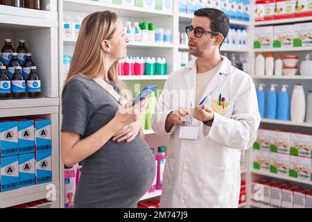 Mann und Frau, Apotheker und schwangere Klientin, die in der Apotheke ein Smartphone benutzen Stockfoto