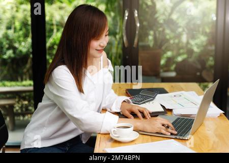 Junge asiatische Frau in eleganter Freizeitkleidung, die am Laptop arbeitet, während sie im Heimbüro neben dem Fenster sitzt. Stockfoto