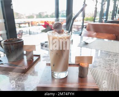 Ein Glas Latte-Kaffee und ein Bärensirup-Eis auf Holzteller mit Sonnenstrahlen im Café und Blick auf den Außenbereich. Stockfoto