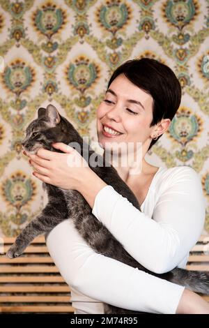 Die junge Frau spielt mit ihrer Katze im Schlafzimmer. Die Frau streichelt die flauschige graue Katze. Stockfoto