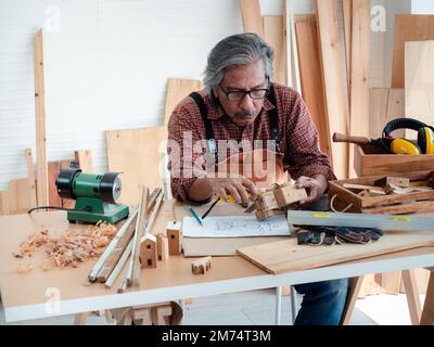 Glücklicher asiatischer Seniorist genießt sein Hobby, indem er in der Werkstatt fleißig Vogelhäuser malt. Stockfoto