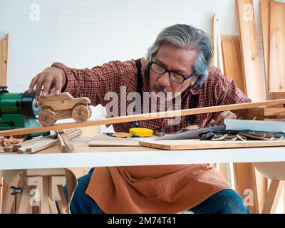 Glücklicher asiatischer Seniorist genießt sein Hobby, indem er in der Werkstatt fleißig Vogelhäuser malt. Stockfoto