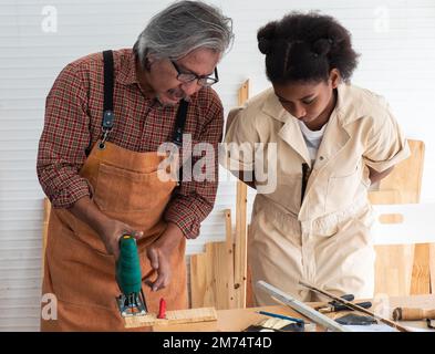 Teenager-Mädchen lernen, eine elektronische Säge zu benutzen und spielen mit ihrem Großvater in der Holzwerkstatt, ein Konzept zur Verbesserung der Entwicklung und des dünnen Designs Stockfoto