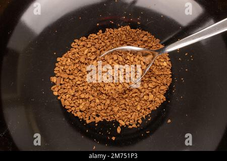 Duftender Instantkaffee auf schwarzem Teller mit Metalllöffel, Makro, Draufsicht. Stockfoto
