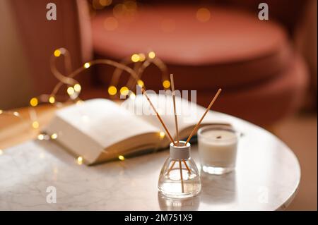 Gemütliche Wohnatmosphäre mit flüssigem Duft in Glasflasche und Stäbchen mit offenem Papierbuch über glühenden Lichtern auf Marmortisch im Wohnzimmer aus nächster Nähe. W Stockfoto