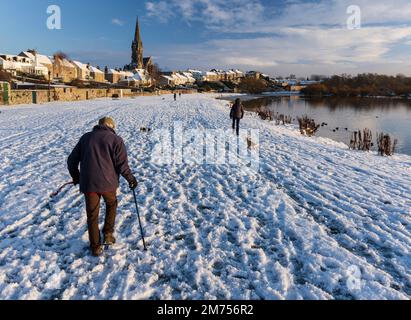 Ein älteres Paar wandelnder Hund im Schnee, kopfsteingepflasterter Fluss, Tweed, Kelso in Schottland. Stockfoto