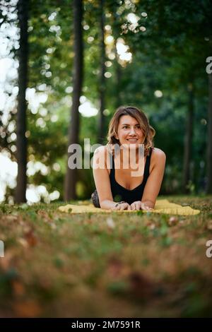 Eine glückliche Frau liegt auf dem Bauch auf einer Yoga-Matte im Wald und macht eine Pause vom Training. Stockfoto