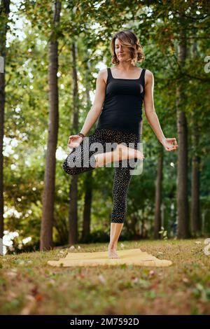 Eine schlanke Frau übt im Herbst Yoga im Wald. Sie ist in der stehenden halben Lotus-Yoga-Pose. Stockfoto