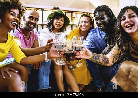 Fröhliche, multiethnische Gruppe erwachsener Freunde, die Spaß beim Feiern mit Rotwein haben - Erwachsene Männer und Frauen, die auf dem Sofa auf Rotwein toasten - Menschen Stockfoto