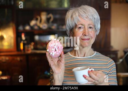 Seniorin trinkt Kaffee und isst einen Donut Stockfoto