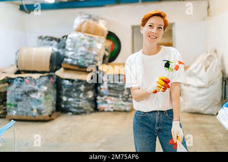 Attraktive rothaarige junge Frau, die freiwillig Handschuhe trägt, Plastikgegenstände in der Hand hält und im privaten Abfallrecycling auf die Kamera schaut Stockfoto