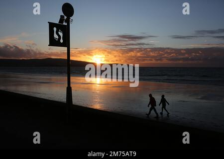 Ayr, Ayrshire, Schottland. Großbritannien Januar 2023. Zwei Silhouette Menschen laufen zügig über nassen Sand auf einem abendlichen Spaziergang am Strand von Ayr entlang. Die Figuren stehen im Kontrast zum Schild, auf dem Fußabdrücke zu sehen sind, die für die schottische lang-Meile werben. Platz für Kopie oder Text. Romantisches Lifestyle-Image Stockfoto