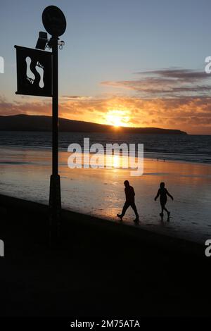 Ayr, Ayrshire, Schottland. Großbritannien Januar 2023. Zwei Silhouette Menschen laufen zügig über nassen Sand auf einem abendlichen Spaziergang am Strand von Ayr entlang. Die Figuren stehen im Kontrast zum Schild, auf dem Fußabdrücke zu sehen sind, die für die schottische lang-Meile werben. Platz für Kopie oder Text. Romantisches Lifestyle-Image Stockfoto