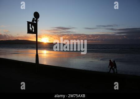 Ayr, Ayrshire, Schottland. Großbritannien Januar 2023. Zwei Silhouette Menschen laufen zügig über nassen Sand auf einem abendlichen Spaziergang am Strand von Ayr entlang. Die Figuren stehen im Kontrast zum Schild, auf dem Fußabdrücke zu sehen sind, die für die schottische lang-Meile werben. Platz für Kopie oder Text. Romantisches Lifestyle-Image Stockfoto