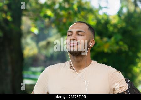 Nahaufnahme Porträt eines Sportlers im Park, eines lateinamerikanischen Mannes, der mit geschlossenen Augen im Park joggt, frische Luft atmet und sich ausruht, mit Kopfhörern joggt und Musik sowie Online-Radio und Podcasts hört. Stockfoto