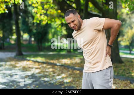 Müde Männer nach dem Joggen im Park, hispanischer Mann hat starke Schmerzen im Rücken und Muskeln nach der Fitness und massiert seine Seite mit der Hand in der Nähe von Bäumen. Stockfoto