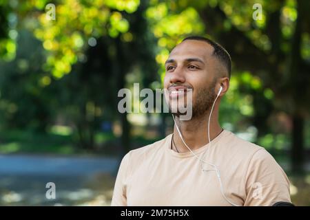 Nahaufnahme eines Mannes im Park, eines lateinamerikanischen Mannes mit Kopfhörern, der Musik und Online-Hörbücher und Podcasts hört, lächelt und wegschaut, joggt und draußen trainiert. Stockfoto