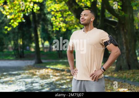Ein lateinamerikanischer Sportler, der sich nach dem Joggen im Park und dem Training ausruhte, schloss die Augen und hörte Musik, Online-Hörbücher und Podcast mit lächelnden Telefonen und Kopfhörern Stockfoto