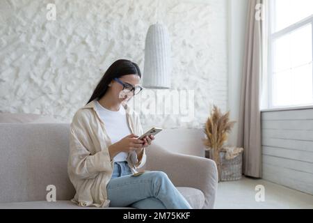 Eine junge, schöne Frau mit Brille sitzt auf einem Sofa am Fenster und benutzt ein Handy, wählt, schreibt, liest Nachrichten, lächelt. Stockfoto